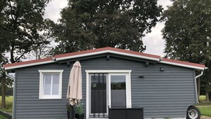 Una piccola casa a un piano con facciata grigia e tetto rosso. Presenta un ingresso con vetro, un piccolo portico e un patio con una sedia da esterno nera e un tavolo.