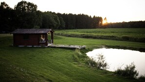 Un piccolo cottage di legno rosso con un portico, adiacente a uno stagno in un prato erboso con alberi sullo sfondo.