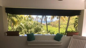 Ein Zimmer mit Fensterblick auf einen üppigen Garten und den Ozean. Eine weiße Bank mit zwei grünen Kissen und ein gestreifter Stuhl sind ebenfalls vorhanden.