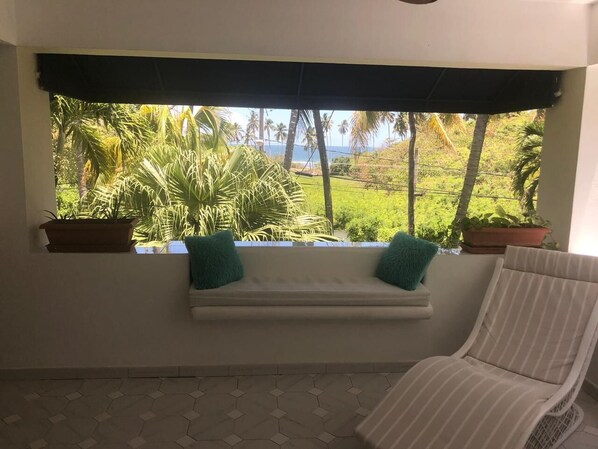 A room with a window overlooking a lush garden and a view of the ocean. There is a white bench with two green cushions and a striped chair.