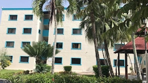 Ein mehrstöckiges Gebäude mit weißer Fassade und blauen Fensterrahmen, umgeben von Palmen und Grünpflanzen.