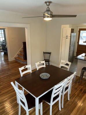 A dining area with a dark wooden table and white chairs, a ceiling fan, and a kitchen with white cabinets and a refrigerator. - Oneonta NY Urban Farmhouse (Oneonta)