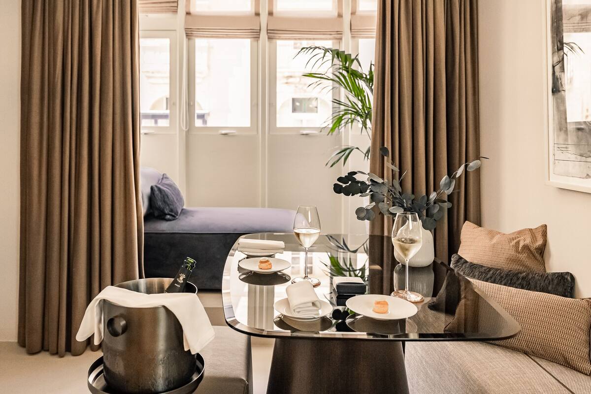 Una sala de estar moderna con una mesa de centro de vidrio, un sofá, una planta en maceta y vistas al exterior a través de amplios ventanales.