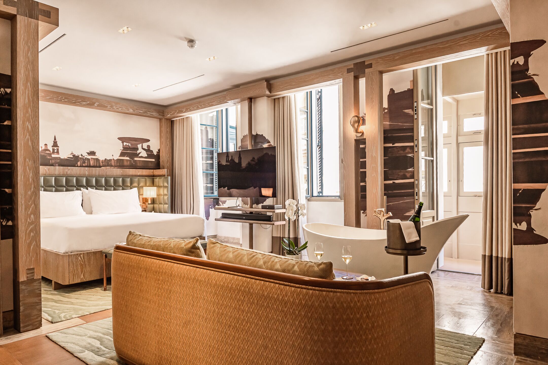 A hotel room with a bed, a sofa, a TV, a dining area, and a view of the cityscape.