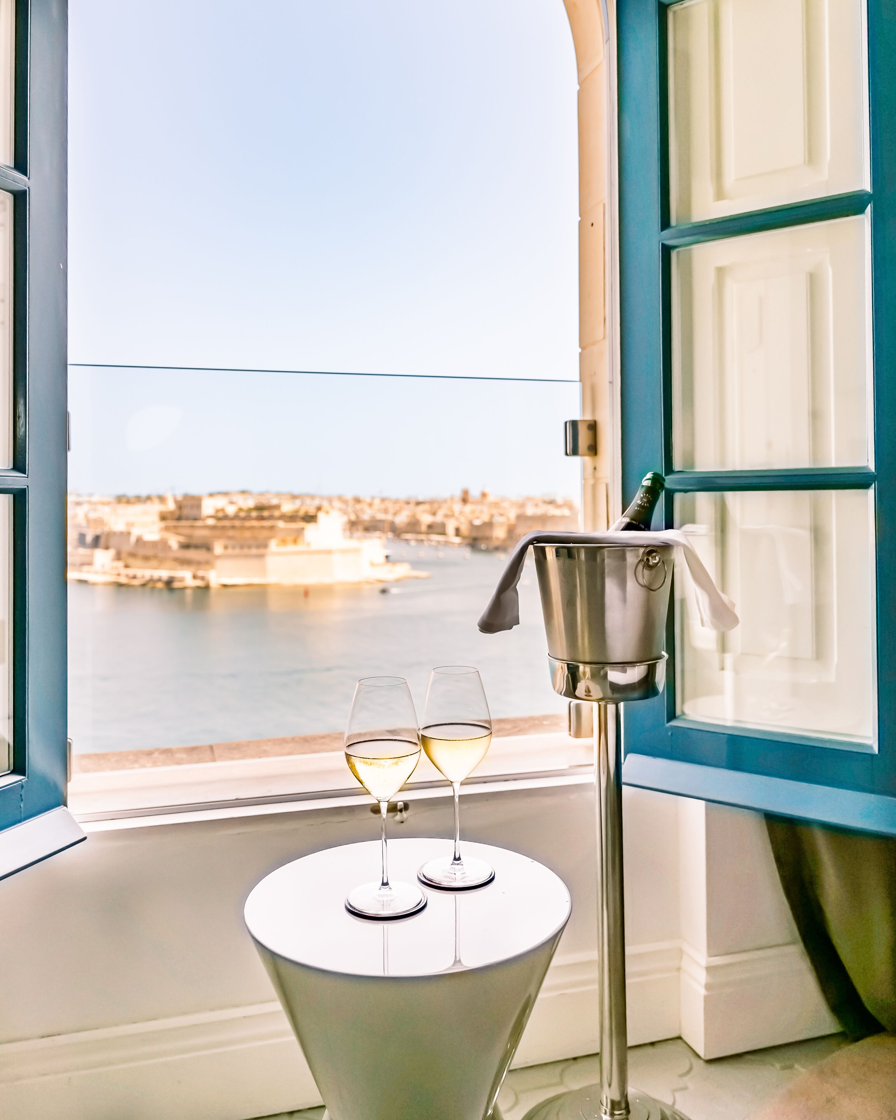A table with two glasses of white wine and a bottle opener near a window overlooking a harbor.