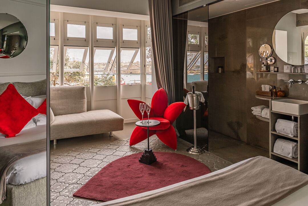 Una habitación de hotel moderna con una cama, una silla roja, una mesita con vasos, un espejo y vistas a un puente y al agua.