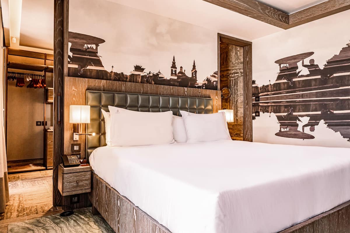 Una cama grande con cabecera de madera, una mesita de noche con lámpara y un mural en la pared con un paisaje urbano.