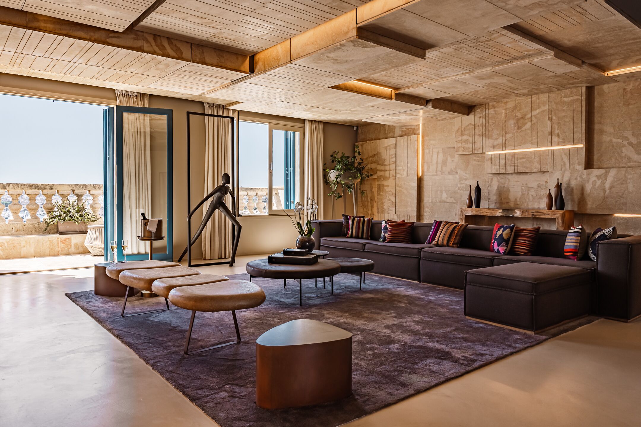 A modern living room with a large sofa, coffee table, and ottoman, a balcony with plants, and a view of the city.