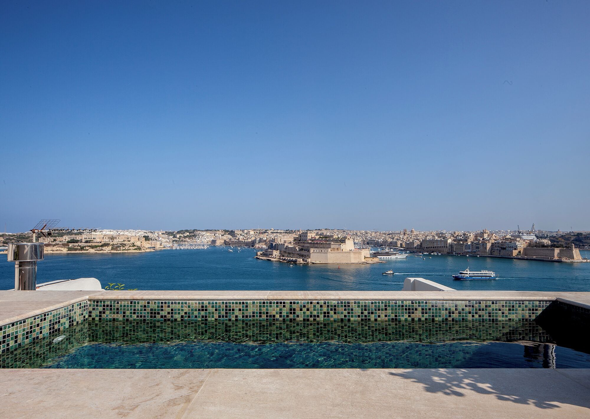 Une piscine bordée de carreaux, une vue dégagée sur le paysage urbain de la côte et une étendue d’eau où naviguent des bateaux.