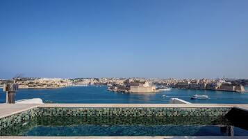 A pool with a tiled edge, a clear view of a coastal cityscape, and a body of water with boats.