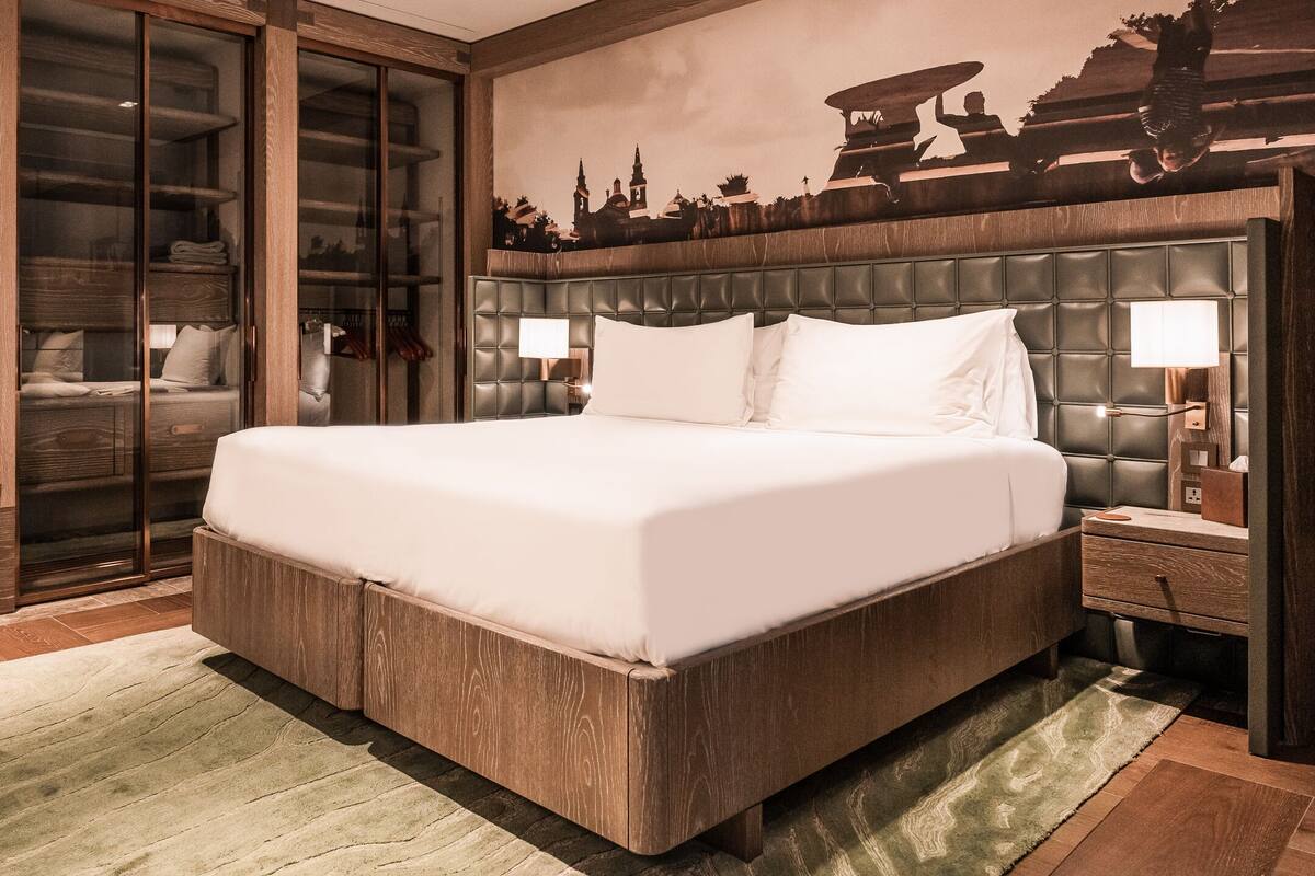 Una cama grande con estructura de madera y cabecera acolchada, una mesita de noche con lámpara, y un espejo grande.