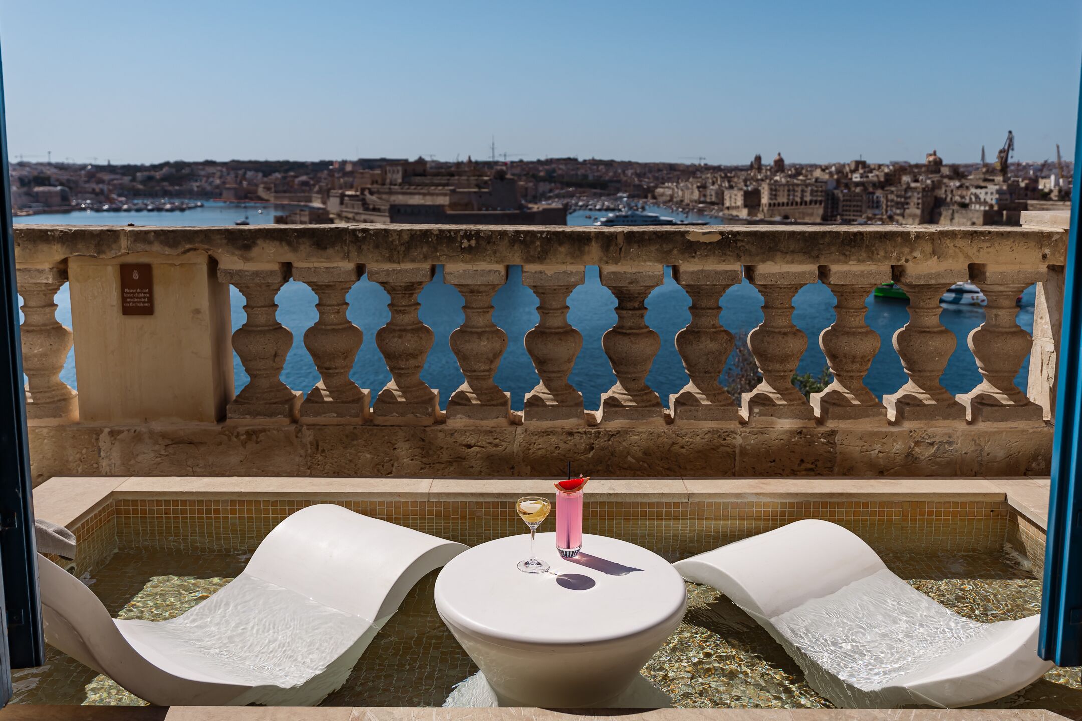 Zona de piscina con dos sillones de descanso, una mesa pequeña con una bebida y vista a un paisaje urbano a través de una baranda decorativa.
