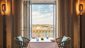 Une table à manger avec deux chaises, une vue sur une ville côtière par une fenêtre ouverte et une balustrade de balcon.