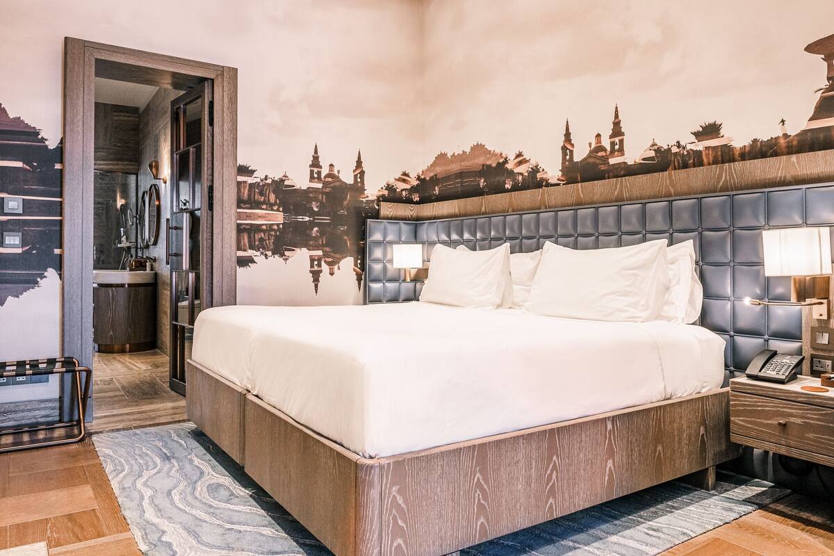 Una habitación de hotel moderna con una cama grande, cabecera con un mural de un paisaje urbano, mesita de noche con lámpara y piso de madera.