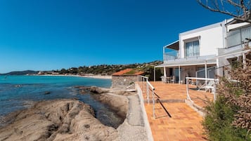 Une propriété située sur la côte, dotée d’un patio donnant sur la mer, d’un mur de soutènement en pierre et d’un bâtiment blanc avec un balcon.