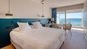 A hotel room with a large bed, a desk with a chair, a ceiling fan, and a view of the sea. - Roc Hotel (Le Lavandou)