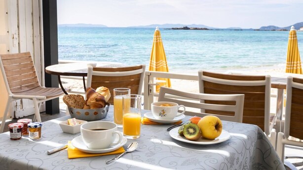 A breakfast setup with croissants, orange juice, coffee, and fruit on a table with a view of the sea and mountains. - Roc Hotel (Le Lavandou)