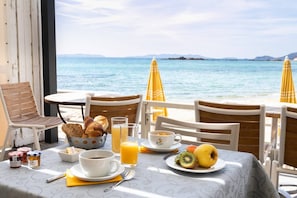 Un plateau de petit-déjeuner comprenant des croissants, du jus d’orange, du café et des fruits, disposé sur une table offrant une vue sur la mer et les montagnes.