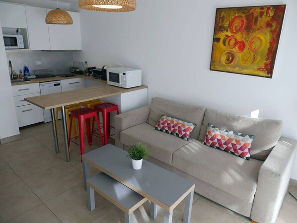 Une cuisine moderne avec des meubles blancs, un four à micro-ondes et un coin repas comprenant une table et des tabourets rouges. On y trouve également un canapé beige avec des coussins à motifs et un tableau accroché au mur.