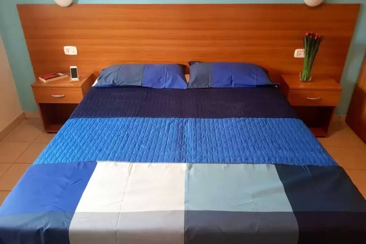 A hotel room with a wooden headboard, a bed with blue and white bedding, two bedside tables with lamps, and a small vase with flowers on one table.