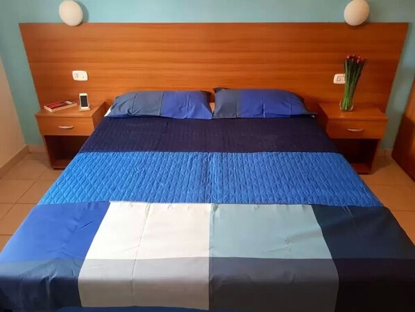 Una camera d'albergo con una testiera in legno, un letto con biancheria azzurra e bianca, due comodini con lampade e un piccolo vaso con fiori su un tavolo.