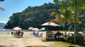 Carrinho de sorvete à beira-mar com uma placa que diz "Coco Gelado" e cadeiras de praia sob guarda-sóis.