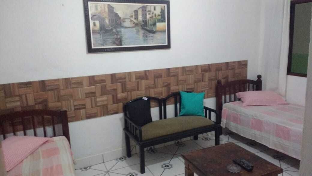 A room with a wooden headboard, a bench with cushions, a bed with a pink blanket, and a small table with a remote control.