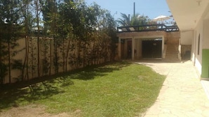 Pátio com caminho pavimentado que leva a uma entrada de edifício, cercado por um muro baixo e vegetação.