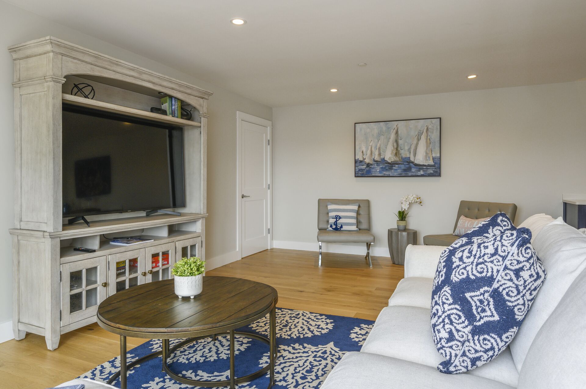 A living room with a large flat-screen TV, a sofa with patterned pillows, a round coffee table, and a painting of sailboats on the wall.