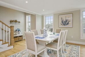 Comedor con una mesa blanca, sillas color beige y un tapete estampado. Se ve una escalera a la izquierda y una ventana con vista a árboles.