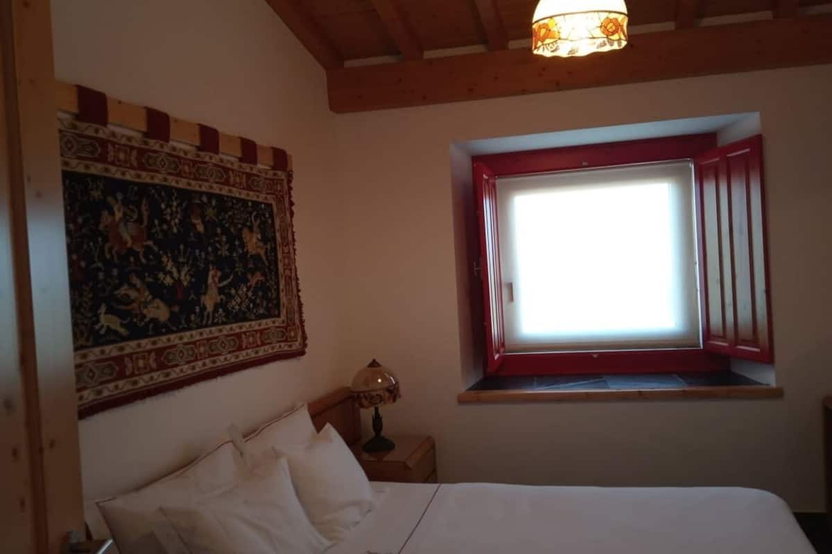Ein Schlafzimmer mit einem Bett, einem Nachttisch mit Lampe, einem Fenster mit roten Rollläden und einem Wandteppich.