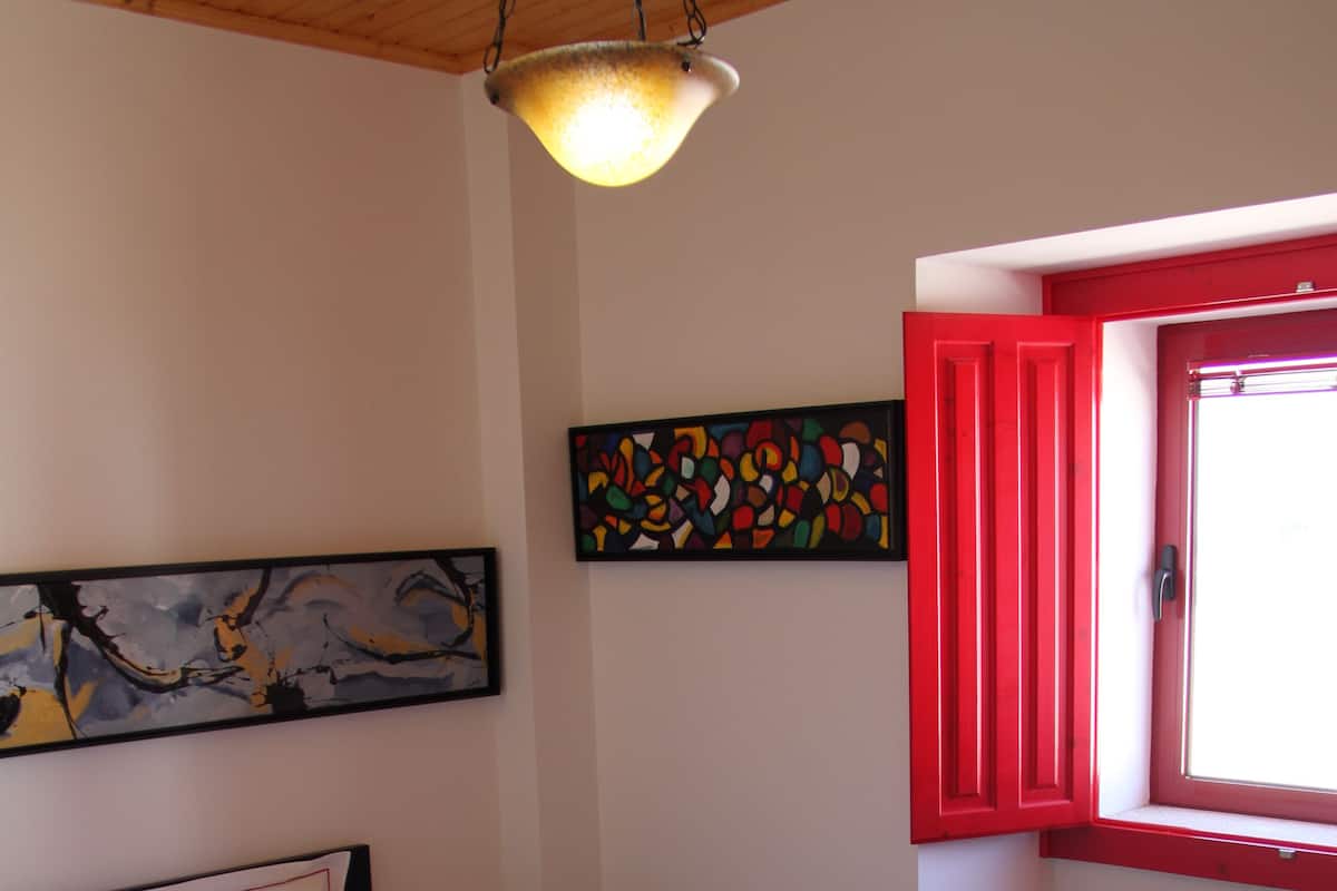 Ein Schlafzimmer mit einem Bett, einem Nachttisch, einem Fenster mit roten Fensterläden und zwei Kunstwerken an der Wand.