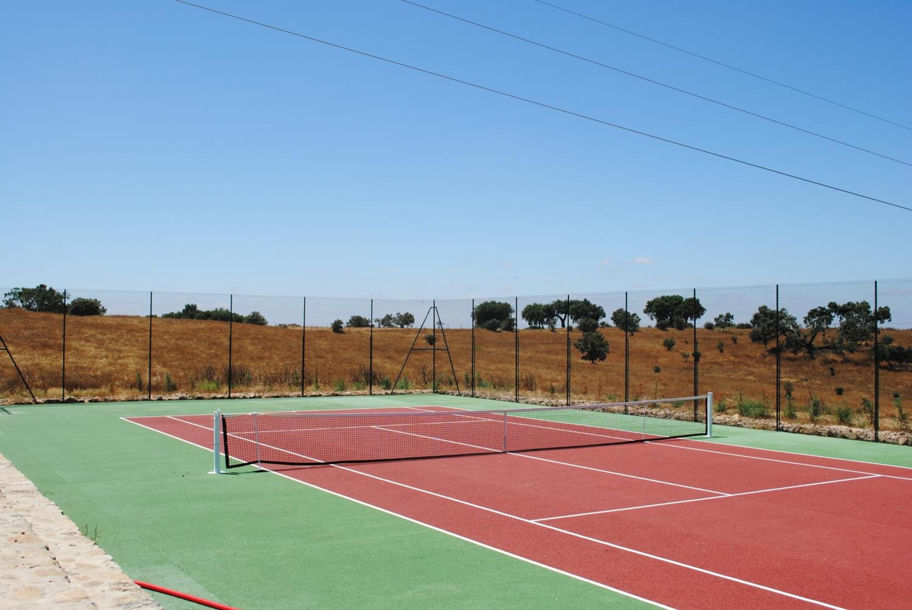 An outdoor tennis court with a net, surrounded by a fence and trees.