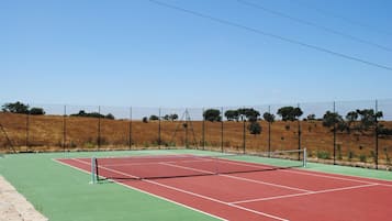 An outdoor tennis court with a net, surrounded by a fence and trees.
