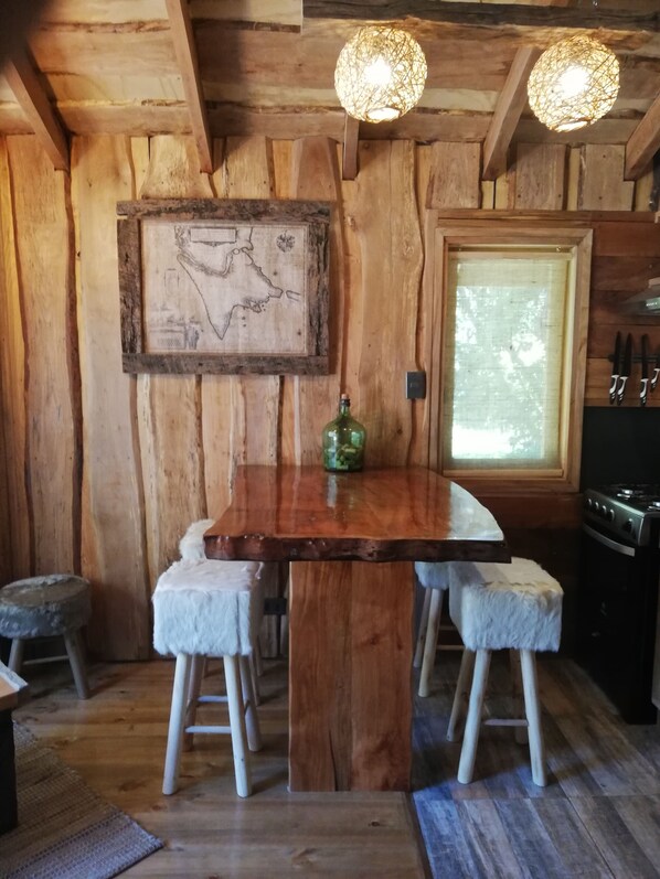 A rustic wooden interior with a wooden dining table, two white cushioned stools, a green bottle, and a map on the wall.