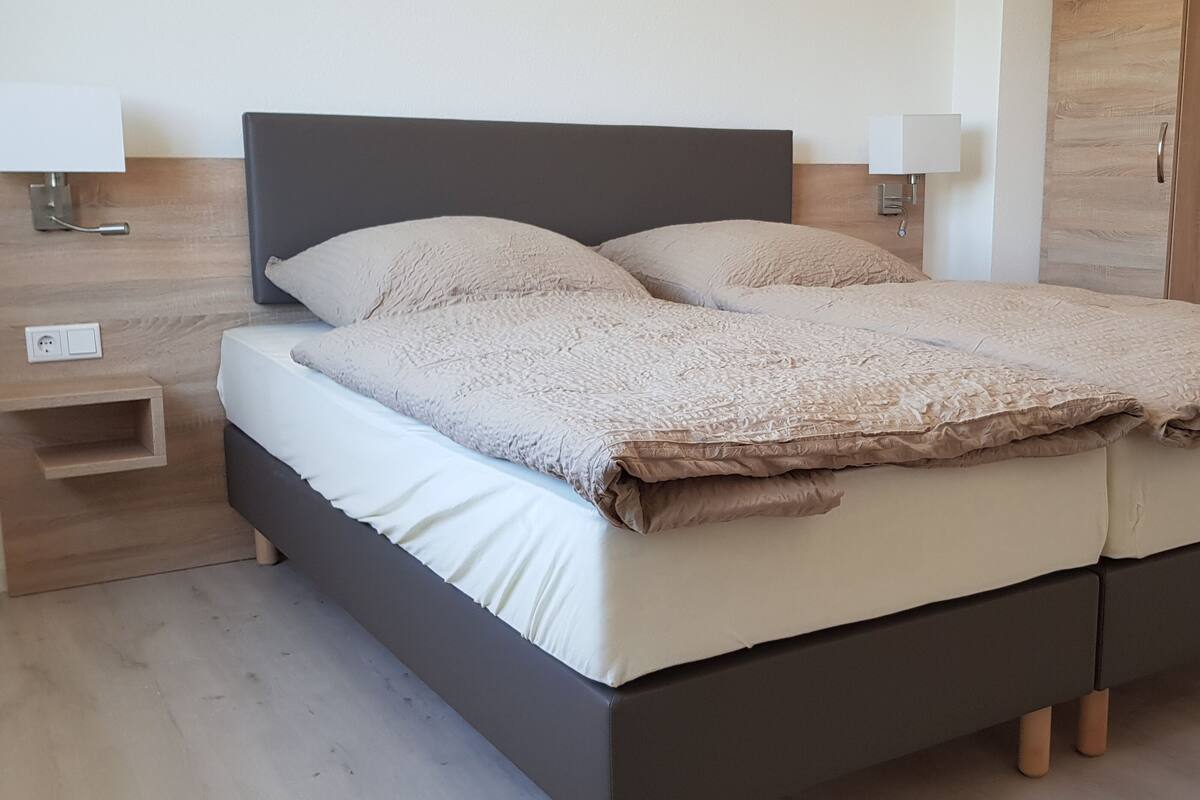 Quarto moderno com cabeceira escura, duas mesas de cabeceira com abajures e um guarda-roupa de madeira.