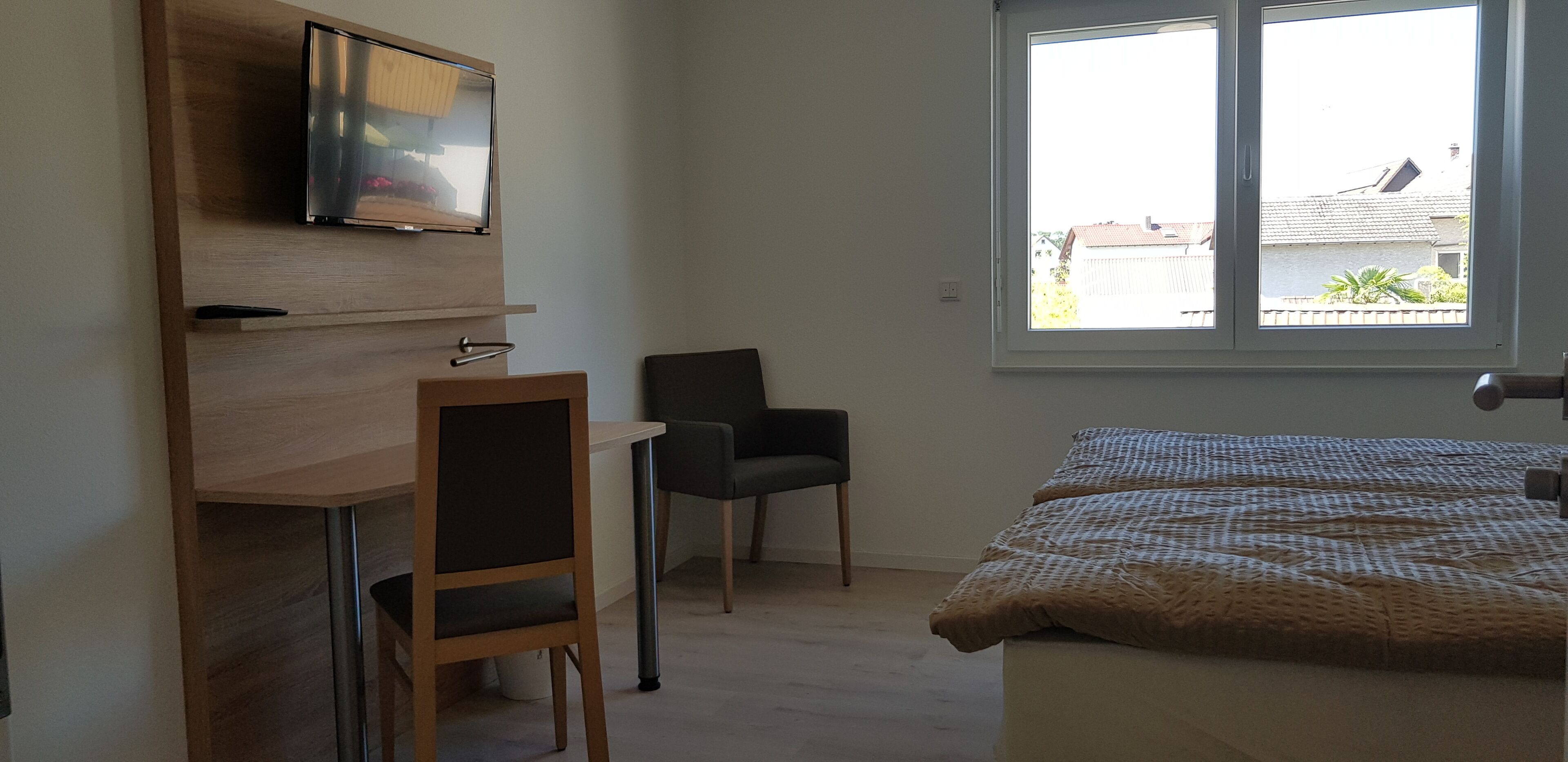 Une chambre à coucher comprenant un lit, un bureau avec une chaise, un téléviseur fixé au mur et une fenêtre donnant sur les bâtiments environnants.