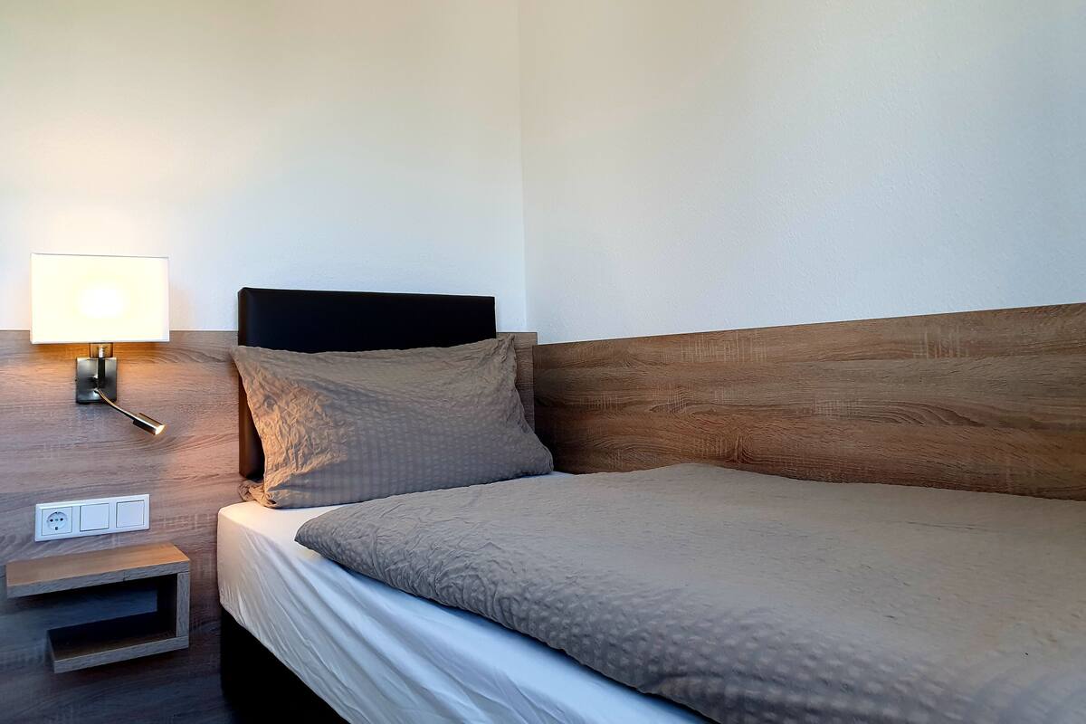 Cama bem arrumada com cabeceira de madeira, mesa de cabeceira com abajur e tomada.