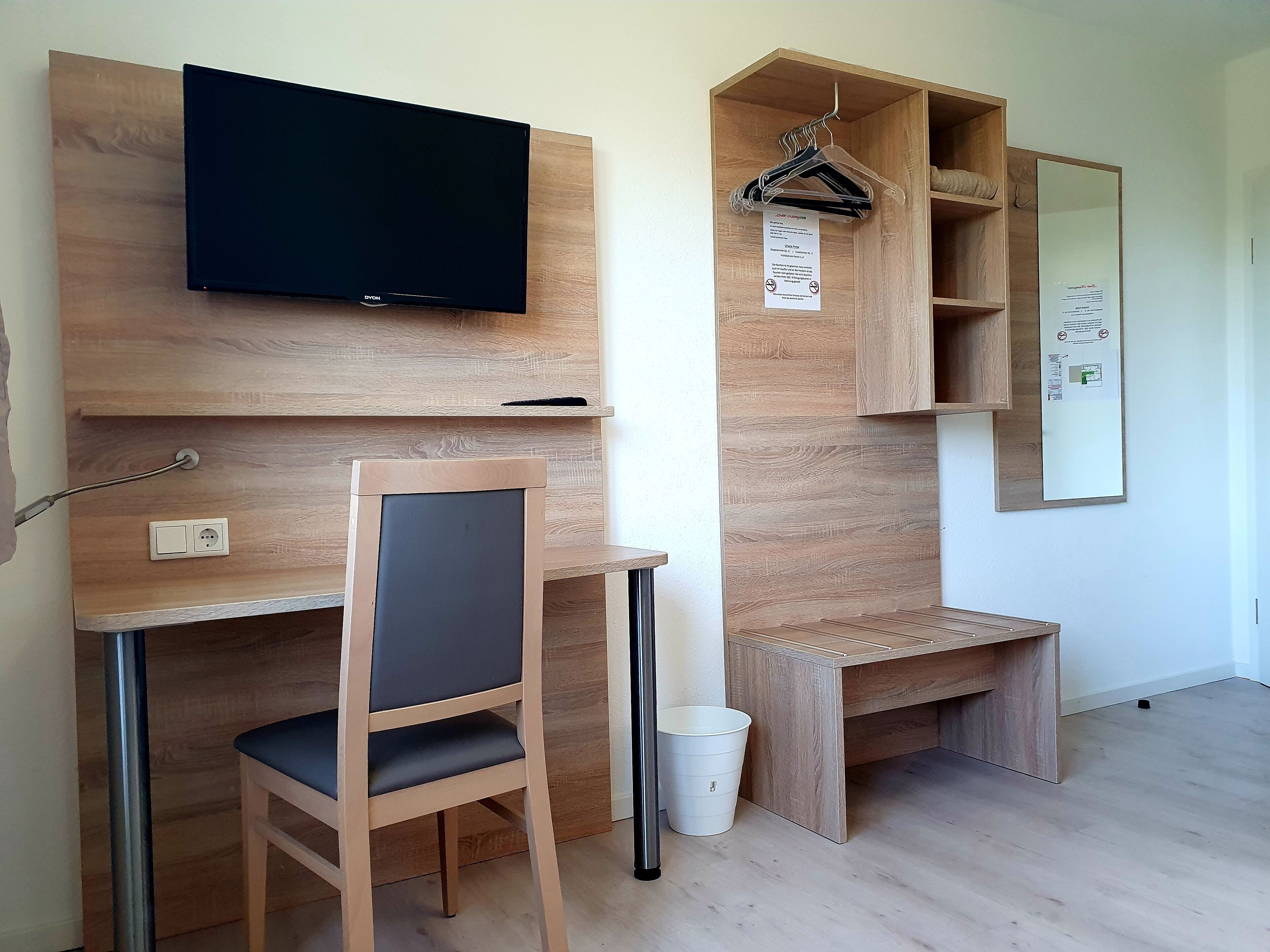 A modern, minimalist room with a wooden desk, a chair, a television mounted on the wall, and a small wooden shelf with a mirror and a hanging coat rack.
