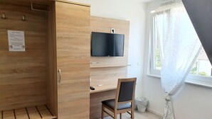 Une pièce de taille modeste, dotée d’une armoire en bois, d’un bureau avec une chaise, d’un téléviseur fixé au mur et d’une fenêtre avec des rideaux.