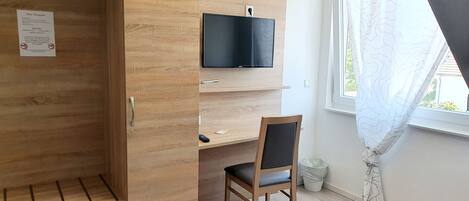 Una habitación funcional con un armario de madera, un escritorio con silla, televisión montada y ventana con cortinas.