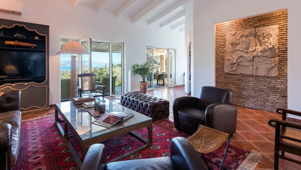 A spacious living room with a glass door leading to a balcony, a large stone wall with a relief sculpture, and a flat-screen TV on a wooden shelf.