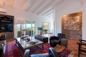 Amplia sala de estar con puerta de cristal que da a un balcón, un gran muro de piedra con un relieve escultórico y un televisor de pantalla plana sobre una estantería de madera.