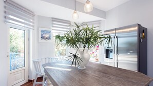 A modern kitchen with a wooden table, white chairs, a large potted plant, and a stainless steel refrigerator.
