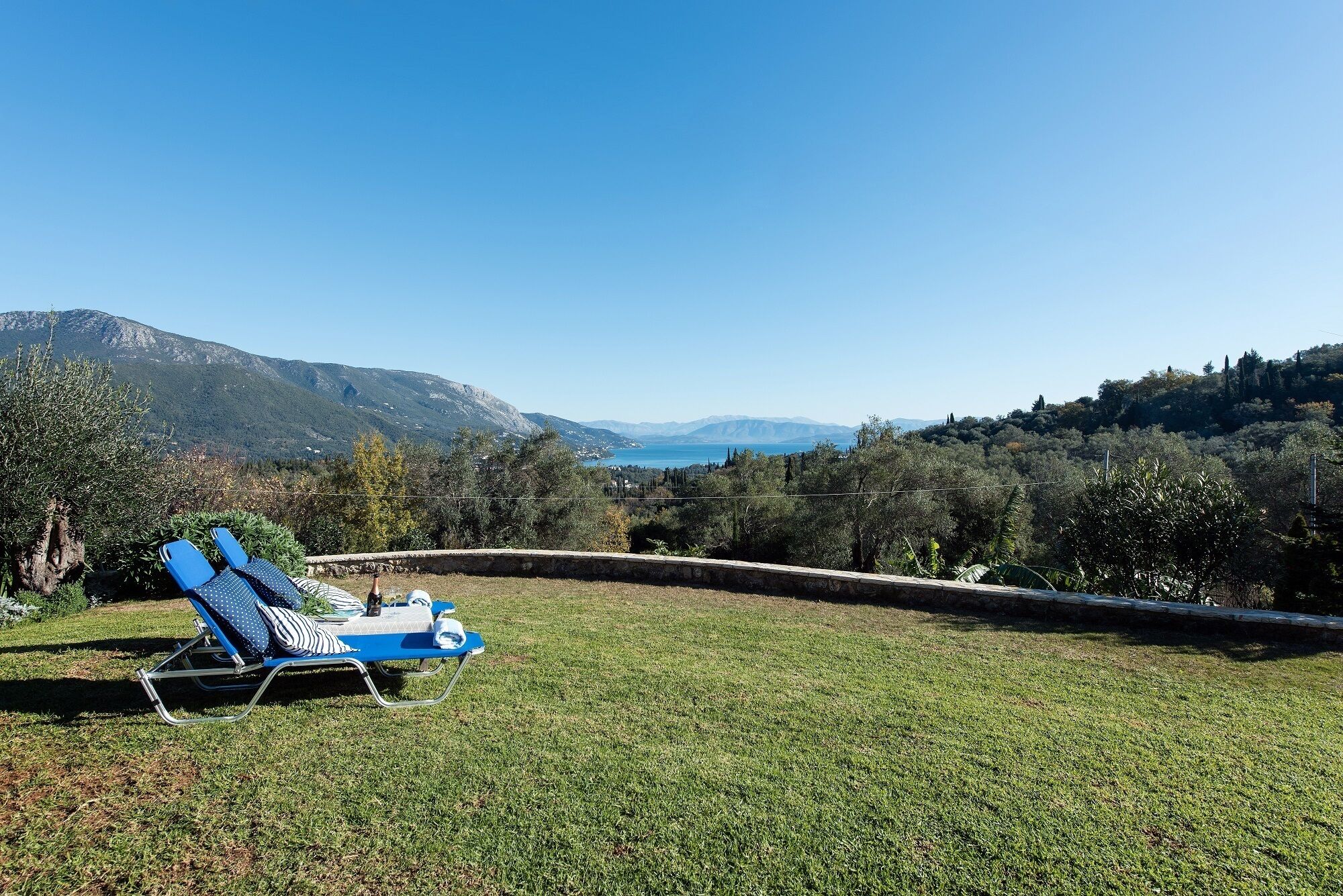 A grassy area with a blue lounge chair and a small table, overlooking a mountainous landscape with a clear sky.