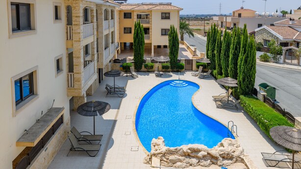 A swimming pool with lounge chairs and umbrellas, surrounded by a building with balconies and a view of the street. - Private Roof Top Terrace Overlooking Pool in Pyla (Pyla)