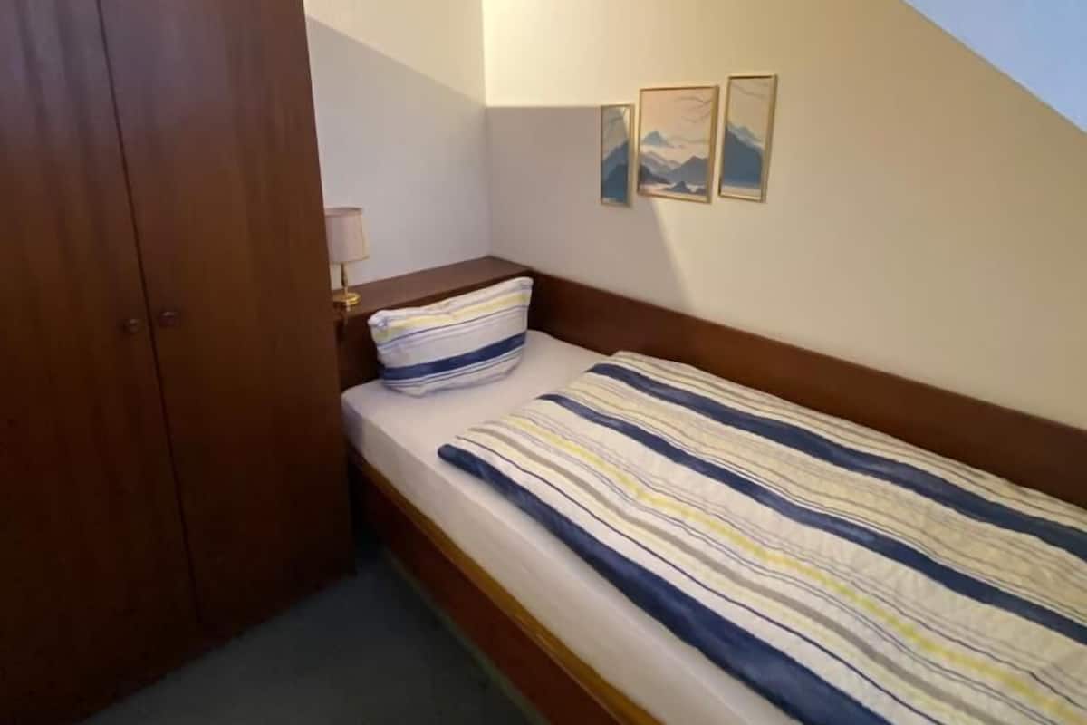 Een kleine kamer met een enkel bed, een schuine plafond, een houten garderobe en twee ingelijste foto's aan de muur.