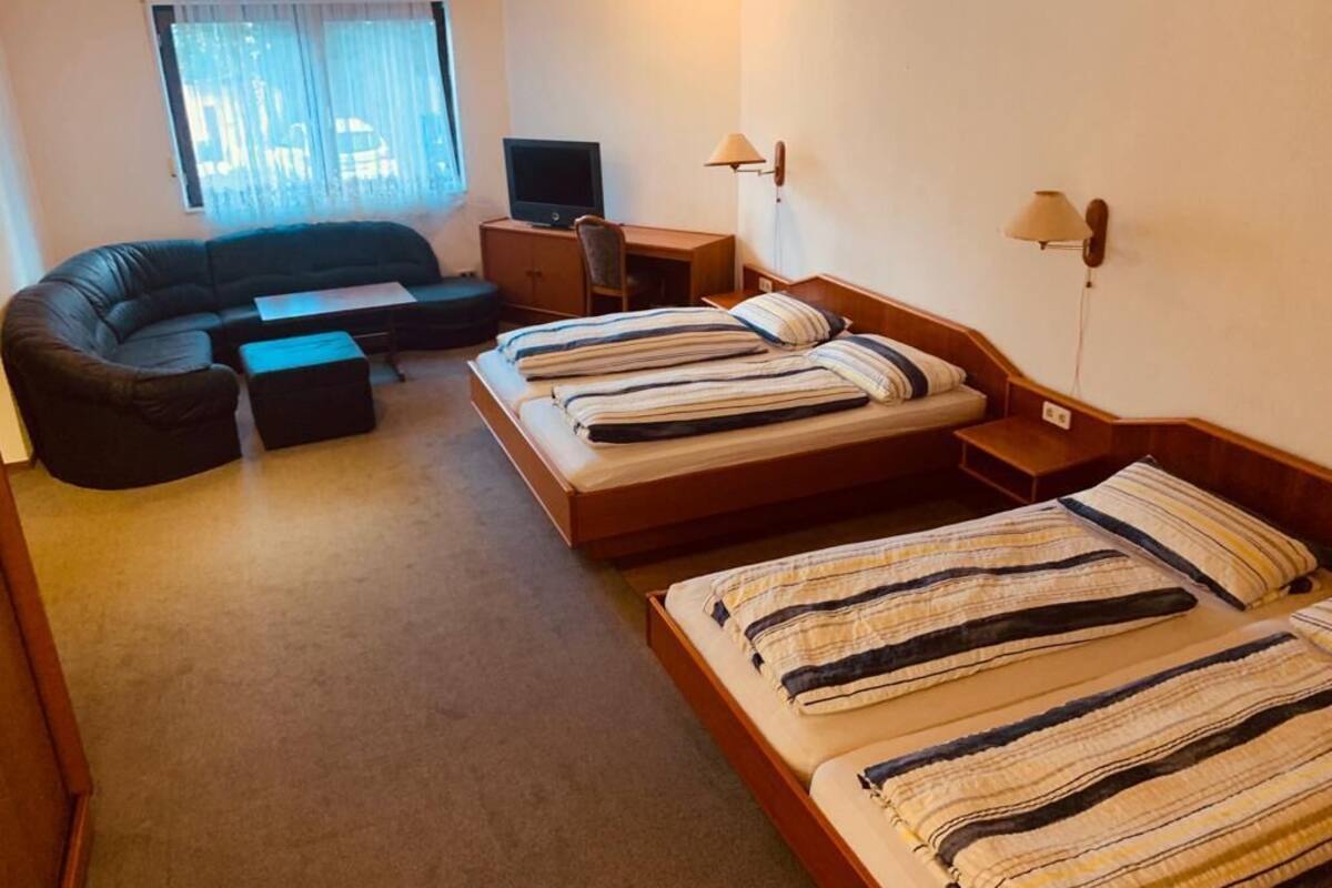 Hotelkamer met een bank, een bureau met een computer, twee bedden met gestreepte lakens en een raam met gordijnen.