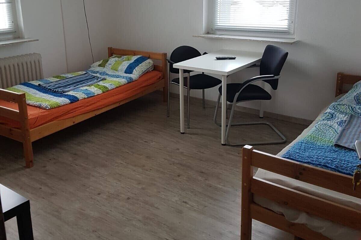 A dormitory room with two single beds, a desk, and a chair.