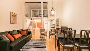 Una sala de estar con sofá de cuero negro, cojines naranjas, y un comedor con sillas de madera y una mesa.